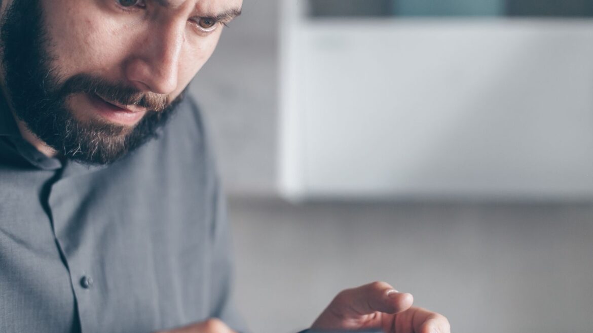Homme barbu en chemise grise, choqué, tenant un smartphone, retrait casino Visa